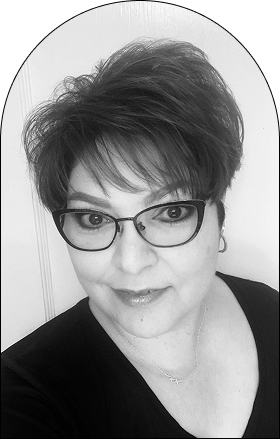 Black and white portrait of a woman with short, dark hair, styled as if fresh from a salon. She wears glasses, earrings, and a necklace, smiling slightly while looking directly at the camera against a light background.