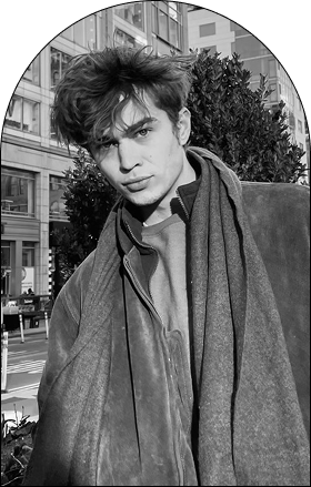 A young man with tousled hair, styled as if fresh from a salon, wearing layered clothing and a scarf stands outdoors in front of a city street and buildings, looking directly at the camera in black and white.