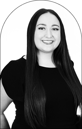 Black and white portrait of a smiling woman with long, straight hair, wearing a short-sleeved, dark top, standing against a plain light background. The image has an oval border at the top—capturing classic Salon style in Corpus Christi.