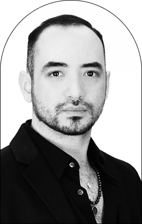 Black and white portrait of a man with stylishly groomed hair and a beard, wearing a dark blazer over a buttoned shirt with a necklace, facing forward with a neutral expression against a light background.