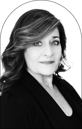 Black and white portrait of a woman with wavy, shoulder-length hair, styled as if ready for the salon, wearing a dark blazer and necklace, looking confidently at the camera against a plain background.