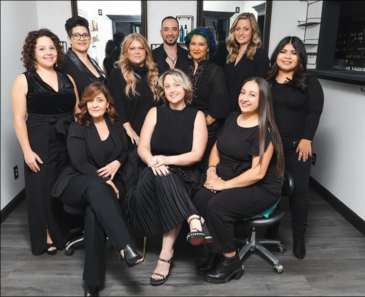A group of ten people, mostly women, pose together in a modern Corpus Christi salon. Most are dressed in black, some sitting and some standing, smiling at the camera amid spa-like design concepts and sleek salon equipment.