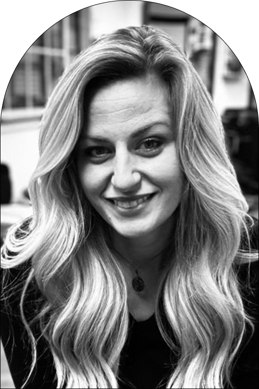 Black and white photo of a woman with long, wavy blonde hair, smiling at the camera. She is wearing a dark top and a pendant necklace, with an indoor Salon background slightly out of focus.