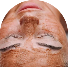 A person lying down with eyes closed, face covered in a brown exfoliating facial mask, enjoying a relaxing spa treatment in Corpus Christi. Their hair is neatly pulled back, suggesting a soothing and rejuvenating skincare experience.