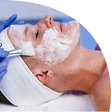A person lies back with eyes closed, wearing a white headband, while a gloved hand applies a white facial mask to their face with a brush—capturing the essence of spa relaxation and hair care.